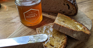 A jar of honey, half a loaf of bread and two slices of thickly cut honey toast and knife on a wooden breadboard