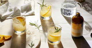 Three glasses of pale liquid, orange slices and sprigs of rosemary sit next to a brown glass bottle and jug of water in afternoon sunshine