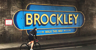 A cyclist rides past a large blue and yellow Brockley mural in the style of old-fashioned station signage