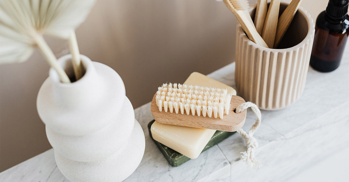 A wooden nail scrubber, soap, a toothbrush pot full of toothbrushes and a vase with dried flower stem on a shelf