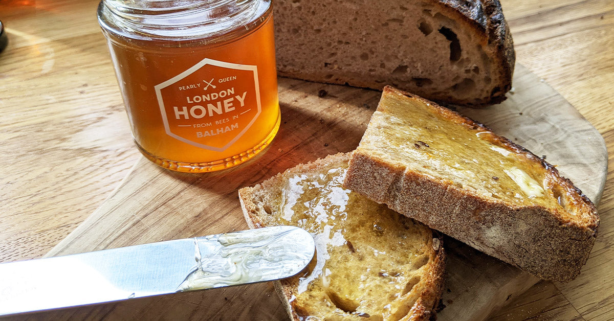 A jar of honey, half a loaf of bread and two slices of honey toast with a knife on a wooden chopping board