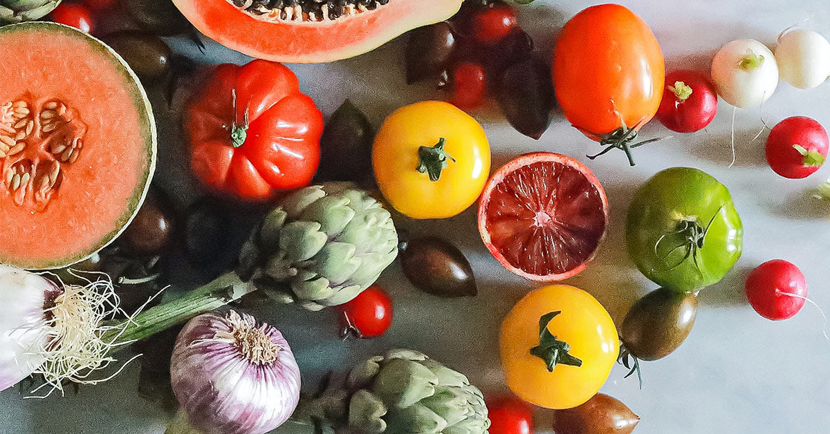 Flatlay of green, red and yellow tomatoes, melons, artichoke and radishes on a white surface